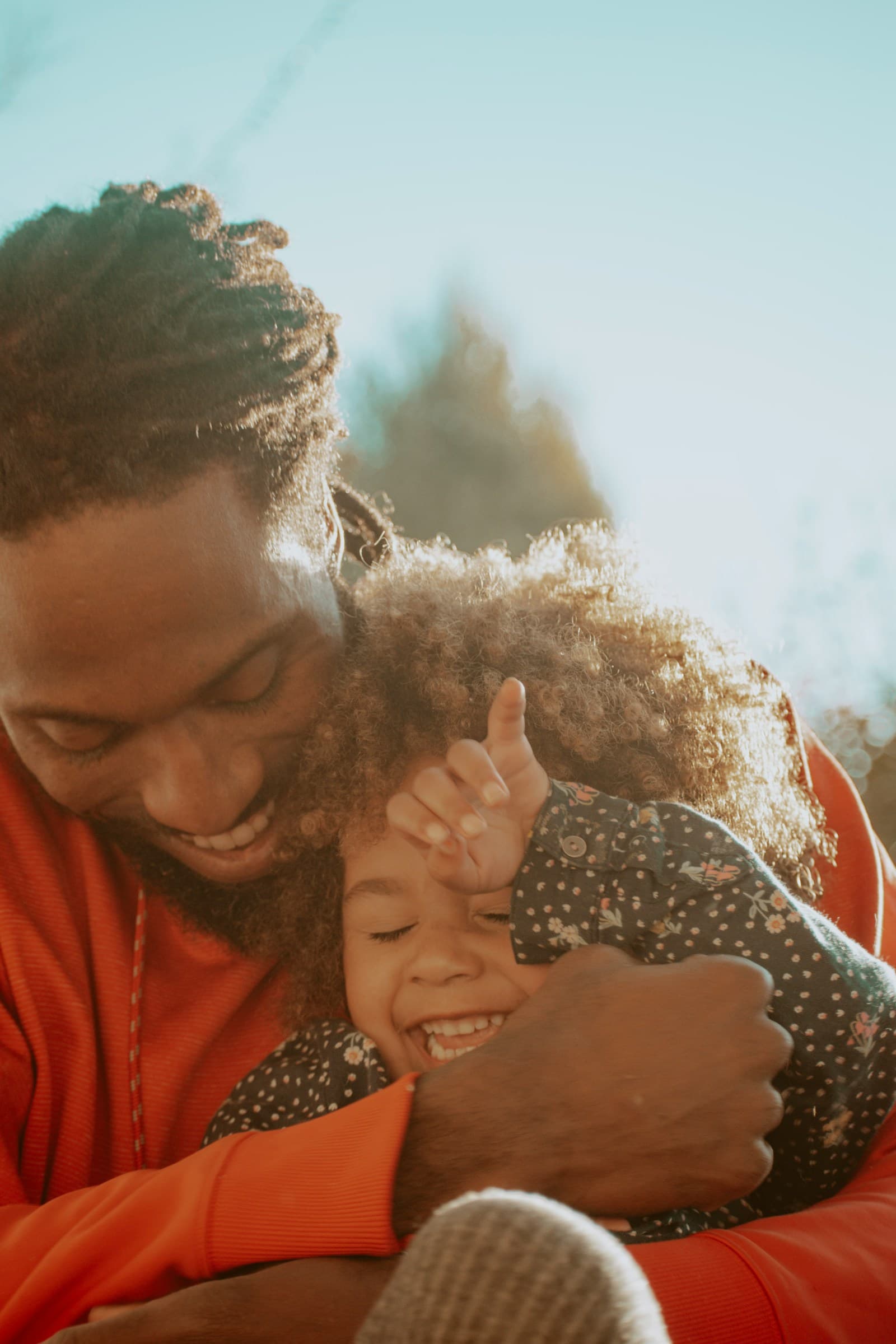 A Black father laughing as he hugs his young daughter in warm sunlight