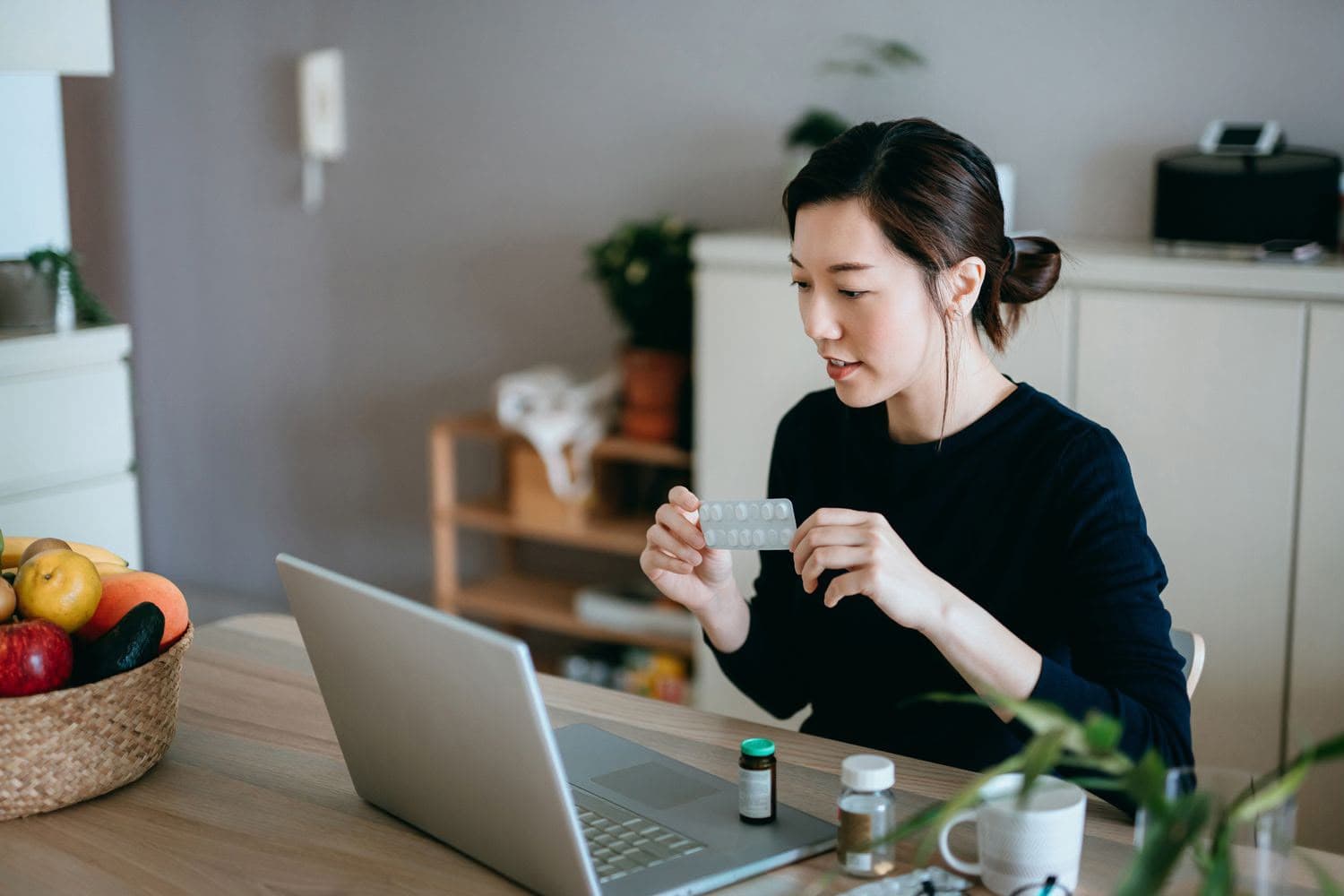 An Asian woman holding up a medication blister pack while talking with her provider on a laptop