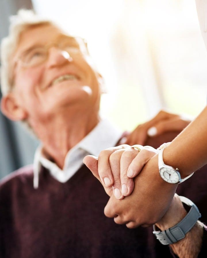 An older man smiling upward as a caregiver's reassuring hand rests on his shoulder, his hand clasped over it