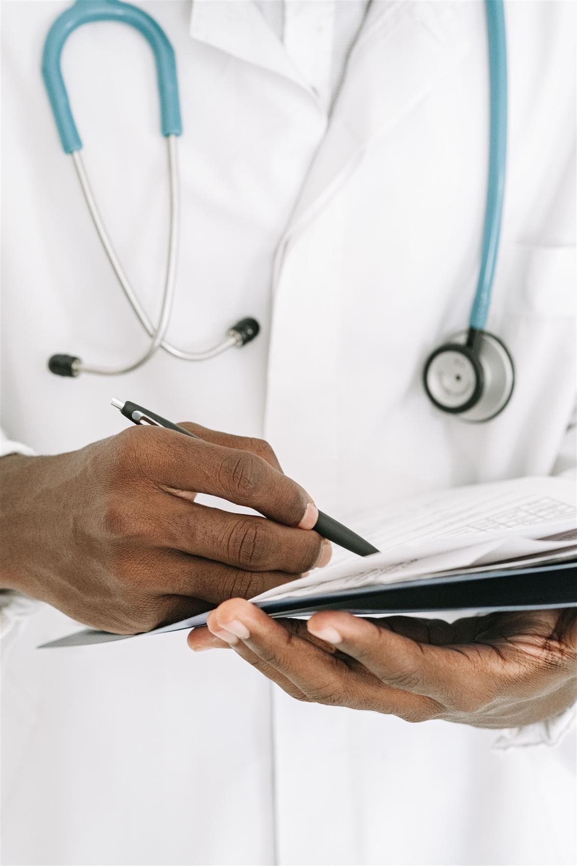 A Black clinician in a white coat writing patient notes on a clipboard