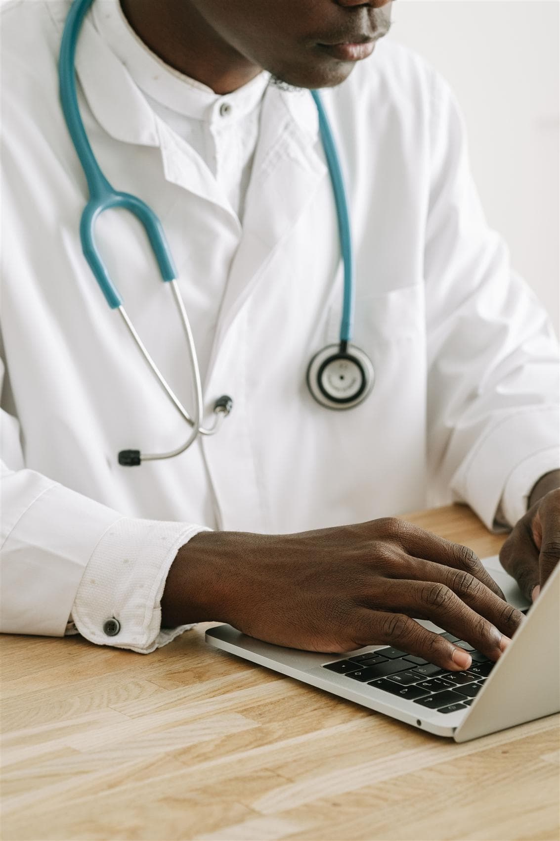 A Black clinician with a teal stethoscope typing on a laptop at a wooden desk